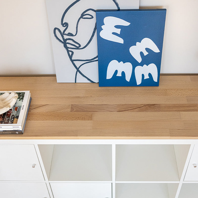 Nahansicht von einer Holzplatte aus Vollholz, die auf einem IKEA Kallax Regal liegt. Auf der Platte stehen Gemälde.