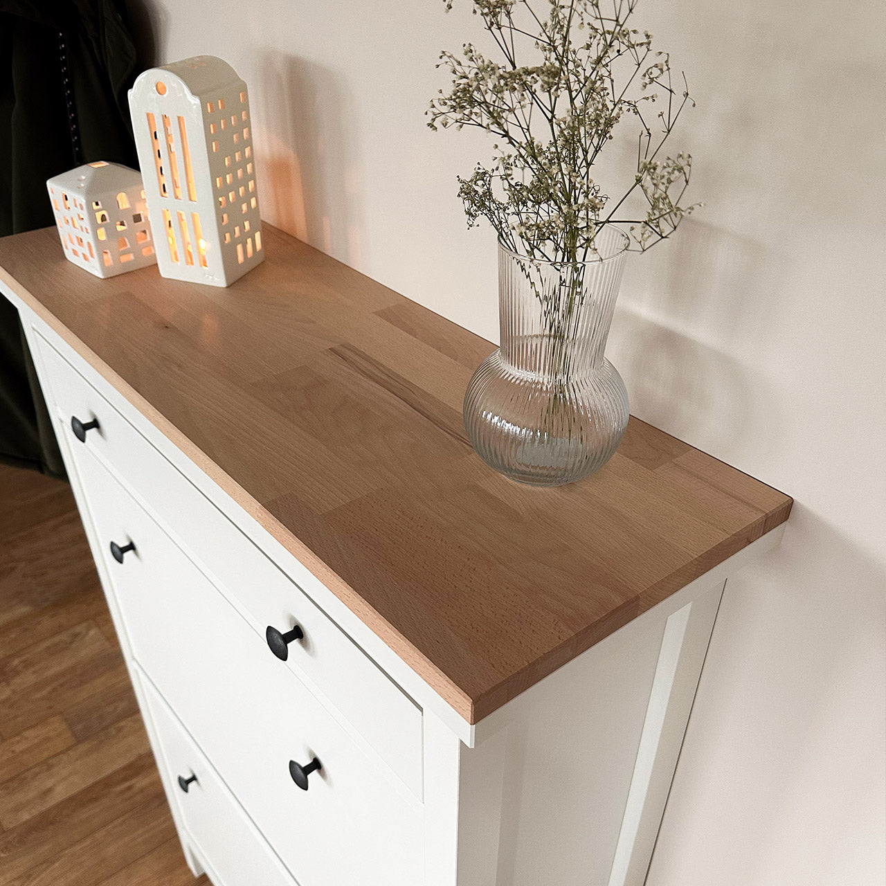 Auf einem weißen Hemnes Schuhschrank von Ikea liegt eine unbehandelte Buchenholz Holzplatte. Auf der Abdeckplatte stehen Deko-Artikel wie eine Vase mit Trockenblumen und Windlichter.